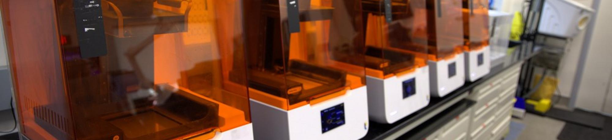 A row of 3D printers with orange-tinted covers sits on a long black countertop in a well-organized lab. The printers are neatly arranged, each featuring a digital display. Shelves above hold labeled storage containers and equipment, while cabinets below provide additional storage. The background includes various tools and workstations, indicating a functional and well-equipped maker space.
